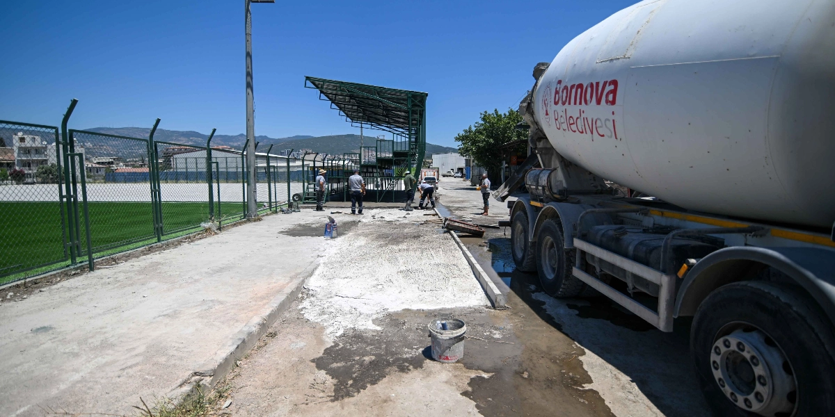 Üzerinde Bornova Belediyesi yazan bir beton mikseri, spor sahasının kenarındaki yürüyüş yoluna beton dökme çalışması yapıyor. İşçiler saha kenarındaki tribünlerin önünde zemin düzenleme ve beton serme işlemlerini sürdürürken, arka planda yeşil çim saha ve dağ manzarası görülüyor.