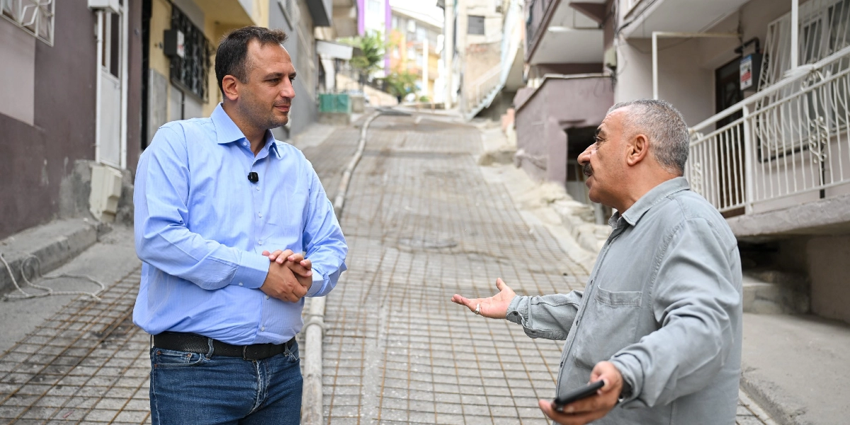 Mavi gömlekli bir yetkili, Bornova'da yenilenmekte olan eğimli bir sokakta, gri gömlekli bir mahalle sakini ile sohbet ediyor. Sokak zemininde beton dökümü öncesi hazırlık aşamasında kullanılan çelik hasır demirler serili durumda.