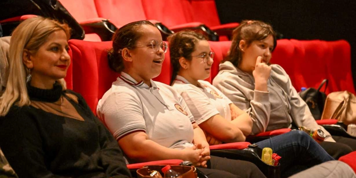 Sinema salonunda kırmızı koltuklarda yan yana oturan, aralarında özel gereksinimli öğrencilerin de bulunduğu bir grup genç kız ve bir yetişkin kadın, ilgiyle karşıya bakarak bir film izliyor.