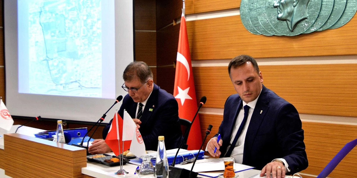 Resmi bir toplantı masasında yan yana oturan, takım elbiseli iki erkek yönetici, önlerindeki evrakları inceliyor. Arka planda Türk bayrağı, duvarda Atatürk kabartması ve üzerinde bir şehir planı veya harita yansıtılan projeksiyon perdesi bulunuyor. Masanın üzerinde mikrofonlar, su şişeleri, küçük Türk bayrakları ve bir bardak çay yer alıyor.