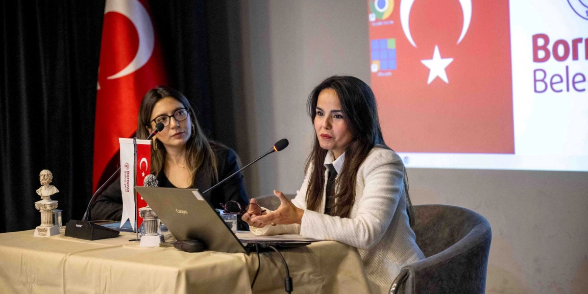 Beyaz ceketli bir kadın, önündeki dizüstü bilgisayarın başında konuşma yaparken, yanında siyah ceketli ve gözlüklü bir kadın onu dinliyor. Masanın üzerinde Bornova Belediyesi logolu küçük bir Türk bayrağı ve küçük bir büst yer alıyor. Arka planda ise Türk bayrağı ve Bornova Belediyesi yazısının olduğu bir projeksiyon yansıtılıyor.