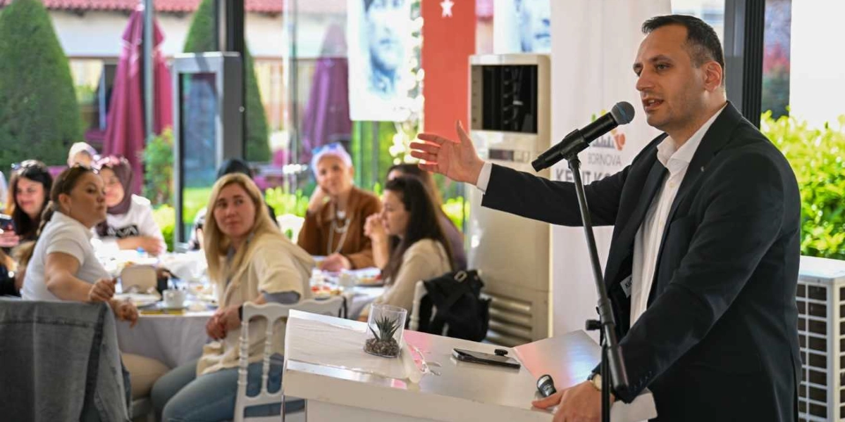 Koyu renk ceketli ve beyaz gömlekli bir erkek, Bornova Kent Konseyi yazılı bir kürsüde mikrofon başında konuşma yapıyor. Sağ elini ileriye doğru uzatarak hitap ettiği dinleyici grubunda, masalarda oturan ve dikkatle dinleyen kadınlar yer alıyor. Arka planda camlı bir mekan ve dışarıdaki yeşil bitkiler görülüyor.
