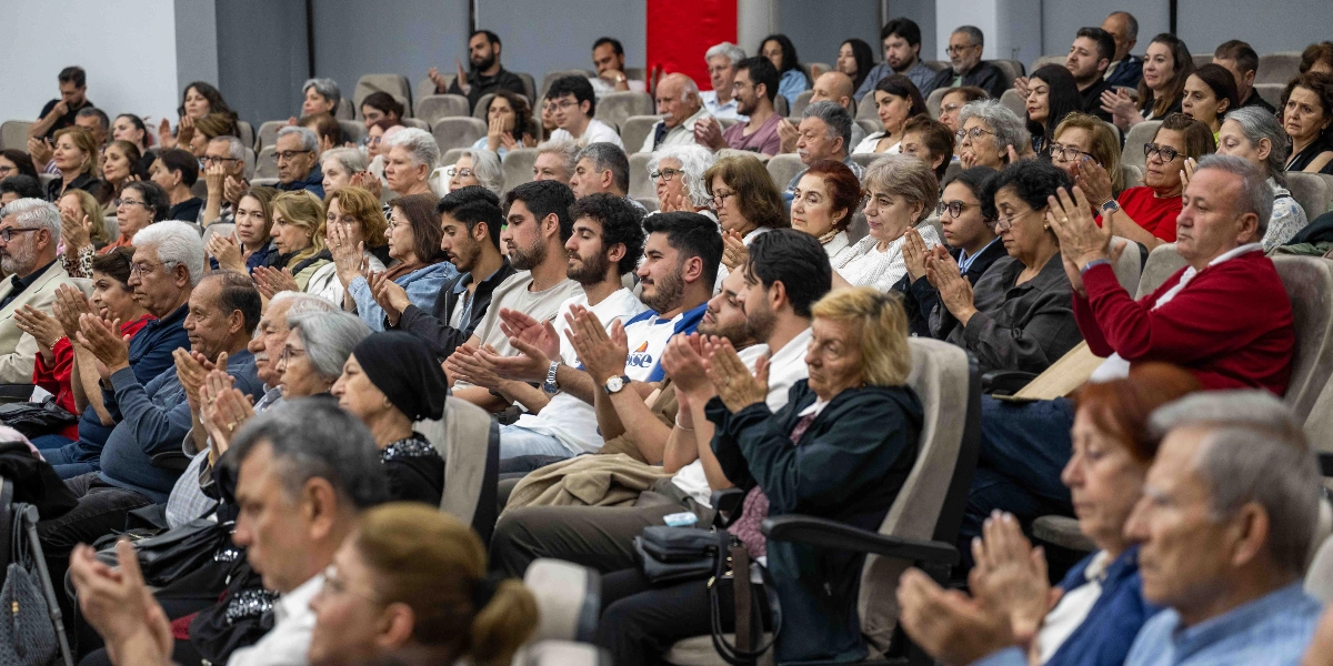 Konferans salonunda oturan, farklı yaş gruplarından oluşan kalabalık bir izleyici kitlesi, sahnedeki bir performansı alkışlıyor. İzleyiciler arasında gençler, orta yaşlılar ve yaşlılar bulunuyor; çoğu kişi ellerini çırparak memnuniyetini ifade ediyor. Salonun oturma düzeni kademeli bir şekilde yükseliyor ve izleyiciler dikkatle sahneye doğru bakıyor.