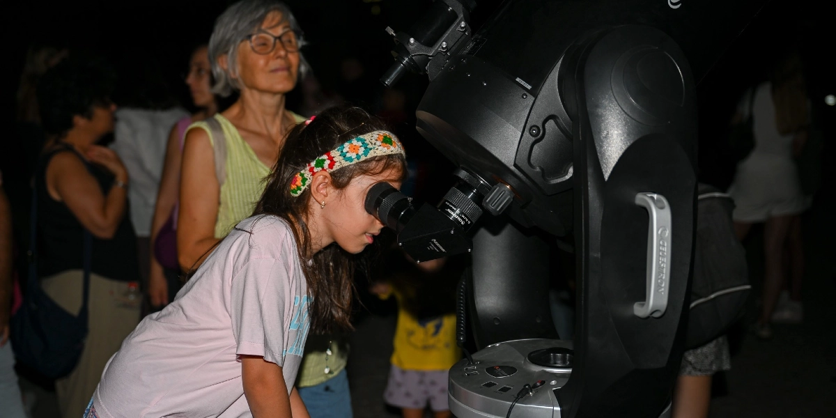 Gece vakti düzenlenen bir etkinlikte, renkli örgü saç bandı takan küçük bir kız çocuğu Celestron marka büyük bir teleskobun merceğinden gökyüzünü inceliyor. Kızın hemen arkasında gözlüklü, gri saçlı bir kadın ve arka planda etkinliğe katılan diğer insanlar yer alıyor.