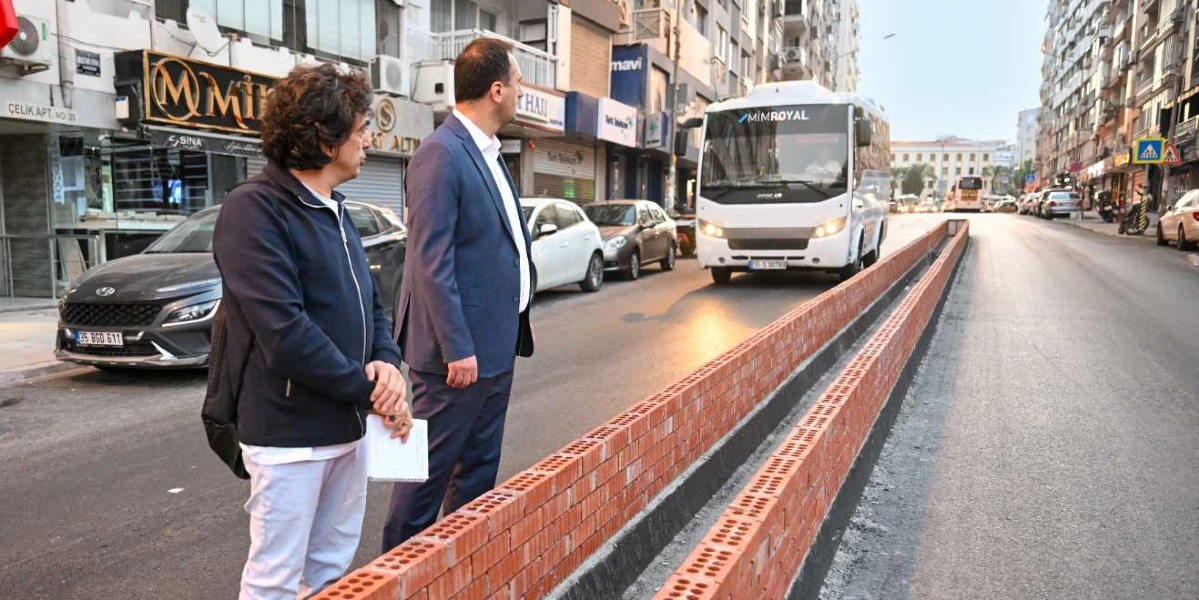 Bornova'da bir cadde kenarında duran, biri koyu renk ceketli ve beyaz pantolonlu, diğeri lacivert takım elbiseli iki erkek, yeni örülmekte olan tuğla bir refüj duvarını inceliyor. Arka planda cadde üzerinde seyir halinde olan beyaz bir servis aracı ve yol kenarında park etmiş otomobiller ile dükkanlar görülüyor.