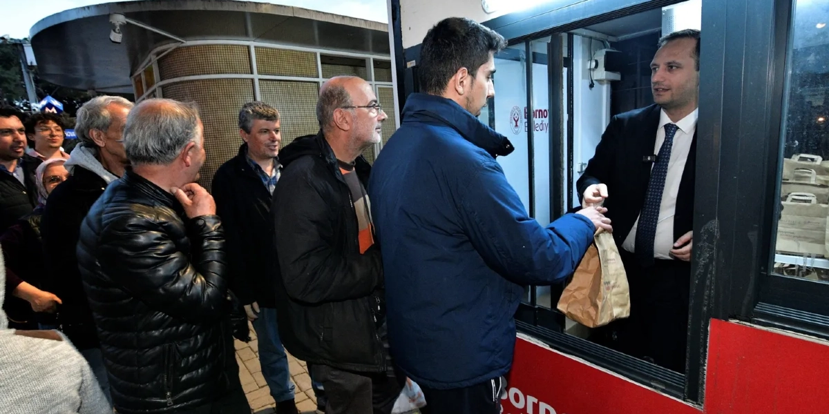 Bornova Belediyesi'ne ait bir stantta, takım elbiseli bir görevli sıradaki vatandaşlara içinde yemek bulunan kağıt paketleri dağıtıyor. Dışarıda bekleyen vatandaşlar sırayla paketlerini alırken, arka planda Bornova Belediyesi logosunun yer aldığı stant yapısı görülüyor.