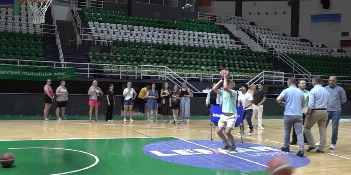 Kapalı bir spor salonunda, basketbol sahasının ortasında duran genç bir erkek, elindeki basketbol topunu potaya doğru fırlatırken görülüyor. Etrafında, etkinliği izleyen farklı yaş ve giyim tarzlarında bir grup genç ve yetişkin bulunuyor. Arka planda ise yeşil ve beyaz koltuklardan oluşan boş tribünler yer alıyor.