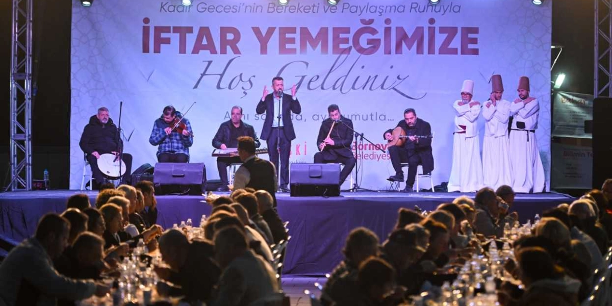 Bornova Belediyesi tarafından düzenlenen iftar etkinliğinde, sahnede bir müzik grubu ve üç semazen yer alıyor. Müzisyenler keman, kanun, ney ve ud eşliğinde performans sergilerken, ortada bir konuşmacı mikrofonla hitap ediyor. Sahnenin arkasındaki büyük pankartta 