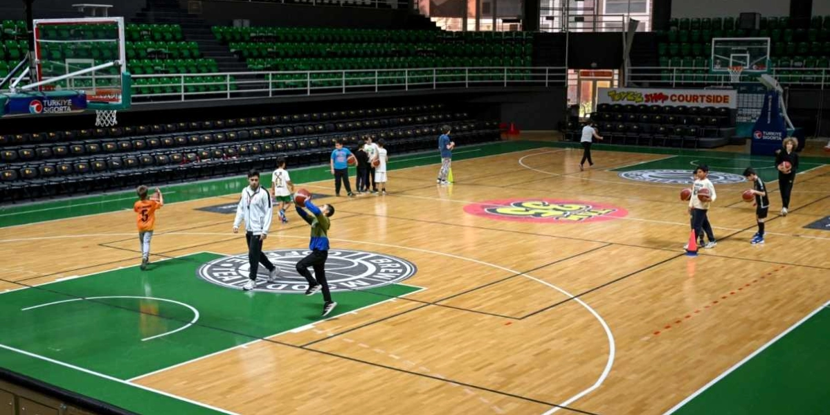 Kapalı bir basketbol sahasında antrenman yapan çocuklar ve onlara eşlik eden bir antrenör. Parke zeminli sahada çocuklar ellerinde basketbol toplarıyla çeşitli hareketler çalışırken, antrenör saha ortasında bir öğrenciye yönlendirme yapıyor. Arka planda boş tribünler ve saha kenarında reklam panoları yer alıyor.