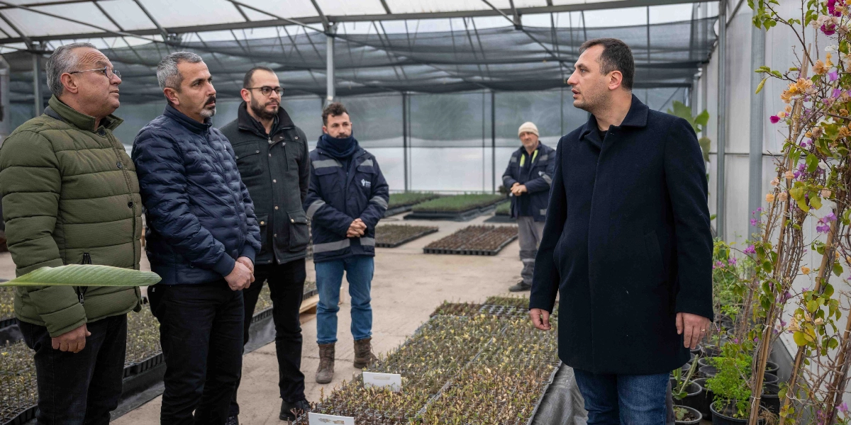 Modern bir serada, Bornova Belediyesi'ne ait olduğu anlaşılan bir tesiste, koyu renkli palto giymiş bir yetkili, yanında bulunan dört kişilik bir gruba bitki üretimi yapılan alanı göstererek bilgi veriyor. Seranın zemininde plastik viyoller içerisinde çok sayıda küçük fide ve saksıda yetişen bitkiler yer alıyor.
