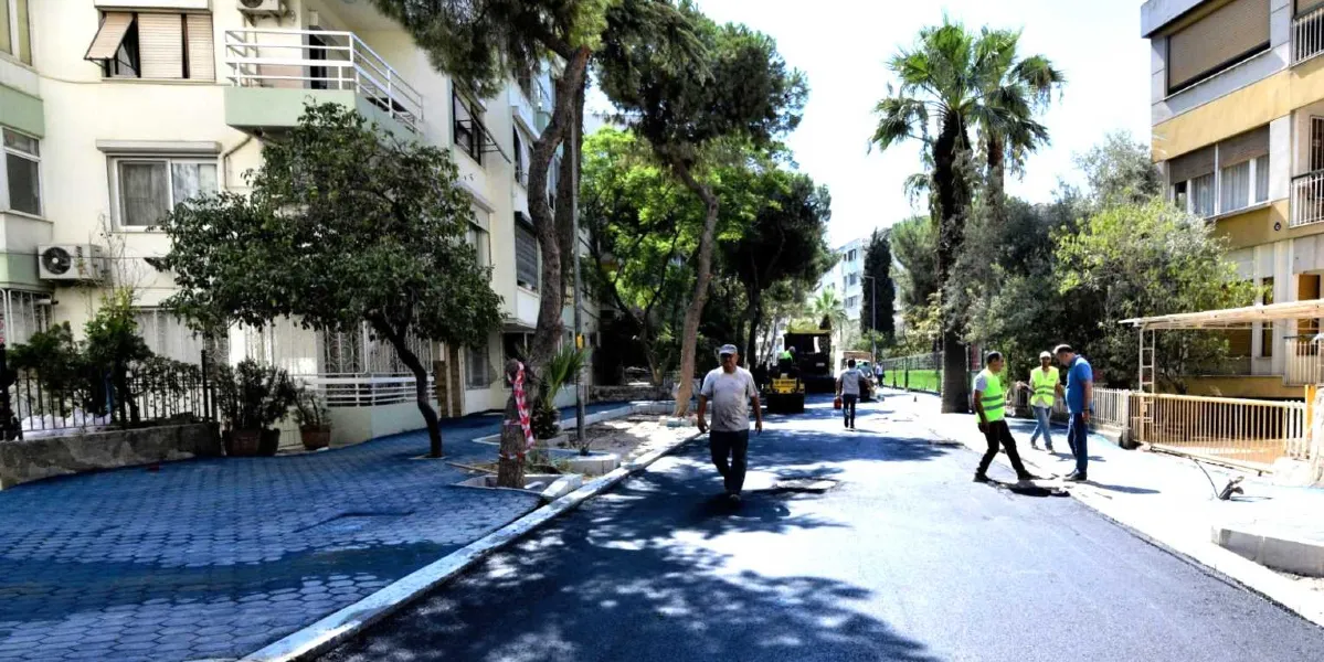 Yenilenmiş asfaltı ve mavi renkli kilit parke taşlı kaldırımlarıyla düzenlenmekte olan bir sokak manzarası. Sokağın ortasında yürüyen bir kişi, sağ tarafta ise yelekli görevliler ve inşaat çalışmaları görülüyor. Çevrede çok katlı apartmanlar, ağaçlar ve palmiyeler yer alıyor.