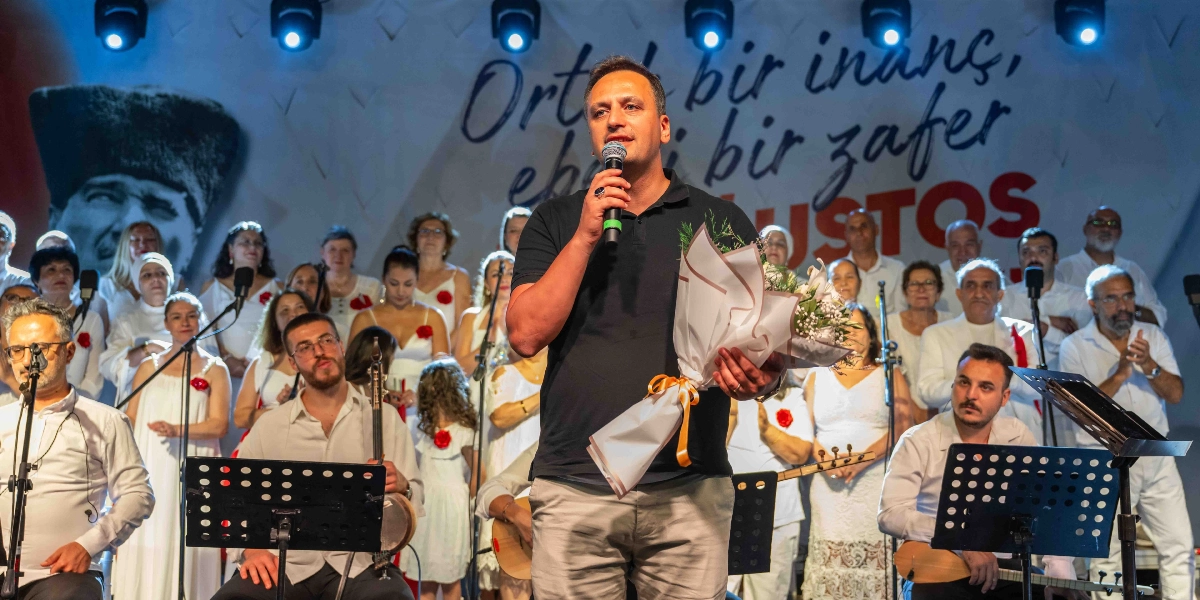 Sahne üzerinde elinde mikrofon ve çiçek buketiyle konuşma yapan bir erkek, arkasında beyaz kıyafetler giymiş kalabalık bir koro ve önlerinde enstrüman çalan müzisyenler bulunuyor. Arka planda Mustafa Kemal Atatürk'ün silüeti ve 