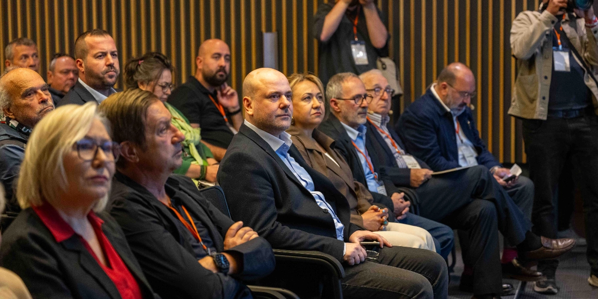 Modern bir konferans salonunda, dikey ahşap panellerle kaplı bir duvarın önünde yan yana oturmuş, dikkatle bir konuşmacıyı dinleyen çeşitli yaş gruplarından katılımcılar. Ön planda sarı saçlı gözlüklü bir kadın, yanında koyu renk ceketli erkekler ve kadınlar yer alıyor. Katılımcıların birçoğunun boyunlarında etkinlik kartları bulunuyor ve sağ tarafta elinde profesyonel bir kamera tutan bir fotoğrafçı ayakta duruyor.