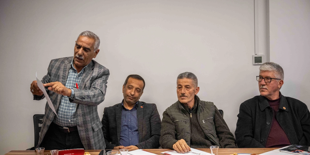 Beyaz bir duvarın önünde, ahşap bir masa etrafında oturan üç kişi ve ayakta durarak elindeki belgeyi gösteren bir kişi yer alıyor. Ayaktaki kişi ekose ceketli, bıyıklı ve elindeki kağıdı işaret ederek konuşuyor. Masada oturanlardan biri lacivert gömlek ve ceket giymiş, diğeri koyu renkli bir yelek ve kazak giymiş, en sağdaki kişi ise gözlüklü, siyah ceketli ve bordo kazaklıdır. Masanın üzerinde çay bardakları ve bazı evraklar bulunuyor.