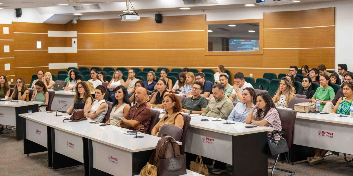 Bornova Belediyesi’nin düzenlediği bir toplantıda, üzerinde Bornova Belediyesi logosu bulunan beyaz masalarda oturan ve dikkatle dinleyen kalabalık bir katılımcı grubu. Ahşap panellerle kaplı modern bir konferans salonunda gerçekleşen etkinlikte, katılımcılar sıralar halinde oturuyor ve önlerinde not defterleri ile su şişeleri bulunuyor.