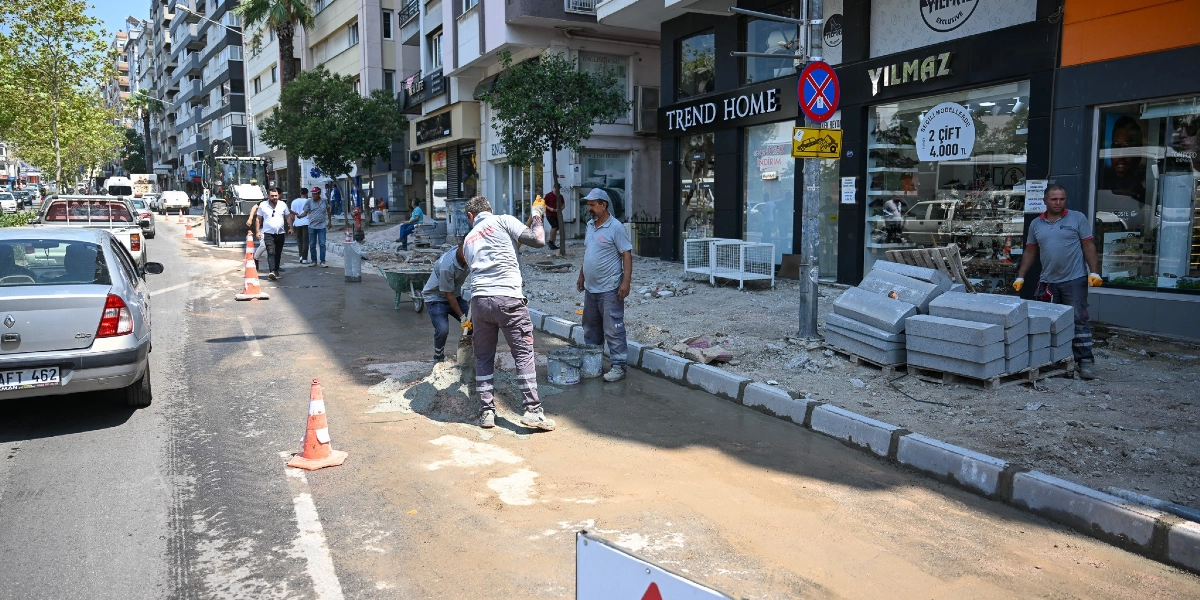 Şehir merkezindeki bir caddede kaldırım yenileme çalışması yapan işçiler görülüyor. Gri tişörtlü üç işçi, yol kenarında harç karıştırıp düzenleme yaparken, bir başka işçi ise palet üzerindeki taşları yerleştiriyor. Çalışma alanı turuncu trafik konileriyle çevrelenmiş durumda. Arka planda dükkanların bulunduğu cadde boyunca park etmiş araçlar ve yaya trafiği yer alıyor. Dükkan vitrinlerinde 