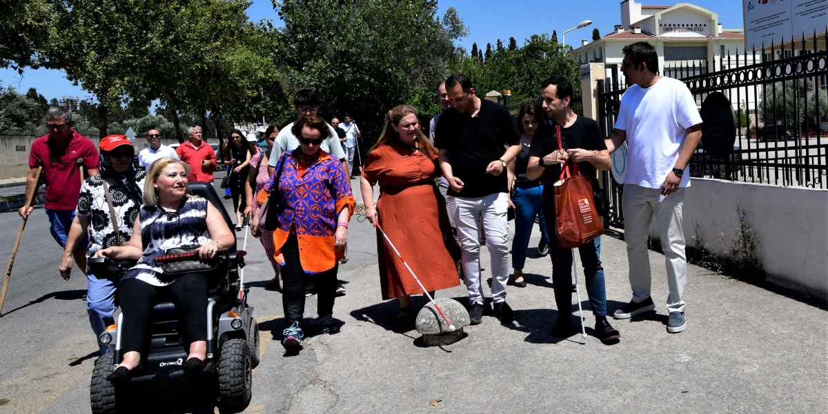 Açık havada, güneşli bir günde kaldırımda yürüyen veya tekerlekli sandalye kullanan engelli bireylerden ve onlara eşlik eden kişilerden oluşan bir grup. En önde, akülü tekerlekli sandalyesinde oturan bir kadın, yanında beyaz baston kullanan turuncu elbiseli bir kadın ve yine beyaz baston kullanan siyah tişörtlü bir erkek yer alıyor. Grubun geri kalanı, aralarında koltuk değneği kullanan bir kişinin de bulunduğu çeşitli katılımcılardan oluşuyor. Arka planda ağaçlar ve İzmir Uygulama Oteli tabelasının bulunduğu bir bina girişi görülüyor.