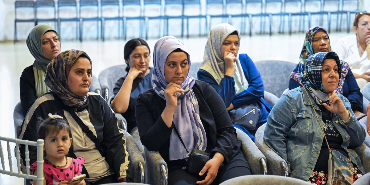 Bir konferans salonunda yan yana dizilmiş gri koltuklarda oturan ve dikkatle dinleyen bir grup kadın. Ön sırada, kucağında küçük bir çocukla oturan kadın, yanında elini çenesine dayamış mor şallı bir kadın ve onun yanında kot ceketli, desenli başörtülü bir başka kadın yer alıyor. Arka sıralarda ise farklı renklerde başörtüleri takmış diğer katılımcılar bulunuyor. Salonun arka planında boş sandalyeler sıralanmış durumda.