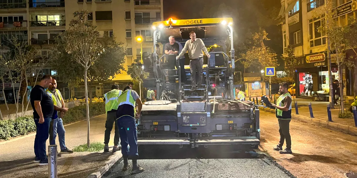 Gece vakti Bornova'da bir caddede asfalt serme çalışması yapılıyor. Sarı ikaz ışıkları yanan Vögele marka asfalt serici makinenin üzerinde ve çevresinde, üzerinde reflektörlü yelekler bulunan bir grup işçi ve yetkili çalışıyor. Makinenin üzerinde iki kişi ayakta dururken, diğer çalışanlar yolun kenarında ve asfalt üzerinde görevlerini sürdürüyor. Arka planda apartmanlar, ağaçlar ve 