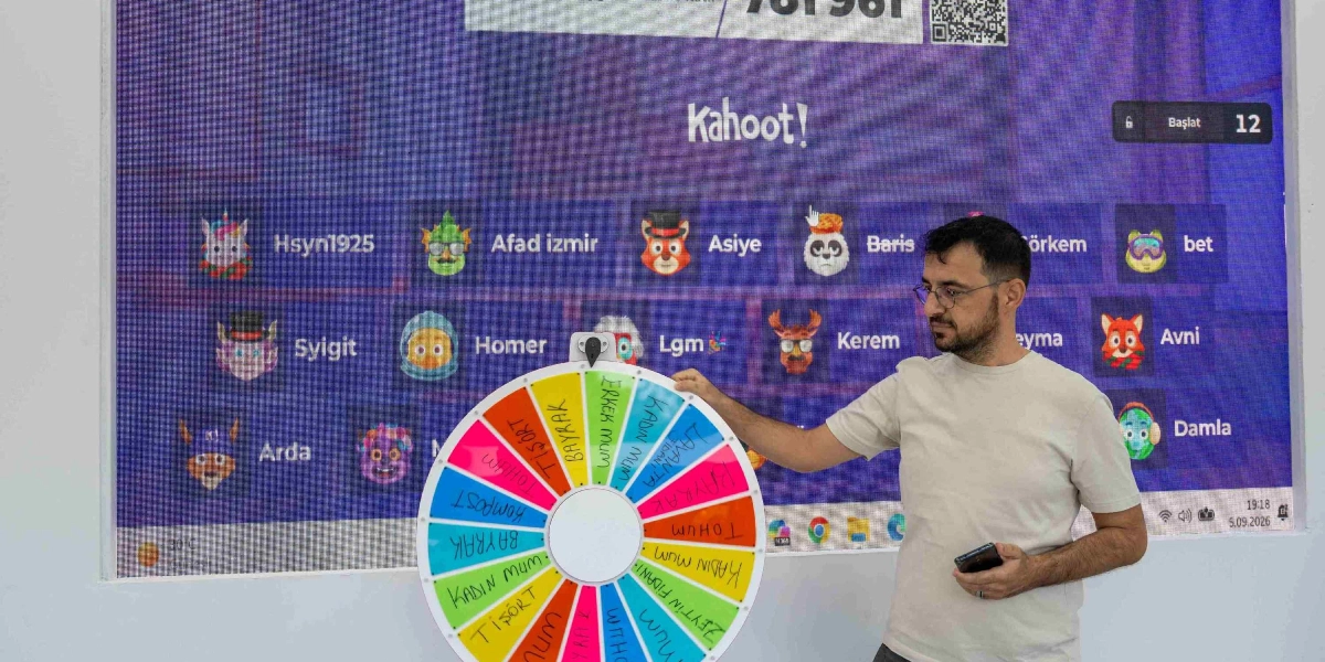 Gözlüklü ve bej tişörtlü bir erkek, arkasında Kahoot oyun ekranının yansıtıldığı büyük bir dijital panonun önünde duruyor. Elinde tuttuğu renkli çarkıfeleğin üzerinde 