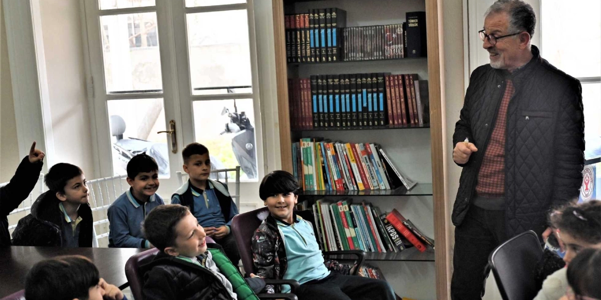 Kütüphanede kitap raflarının önünde duran gözlüklü, orta yaşlı bir erkek, etrafındaki ilkokul çağındaki çocuklara bir şeyler anlatıyor. Çocuklar ilgiyle onu dinlerken, bazıları gülümseyerek etkileşime katılıyor. Arka planda kitaplarla dolu raflar ve dışarıyı gösteren büyük pencereler yer alıyor.