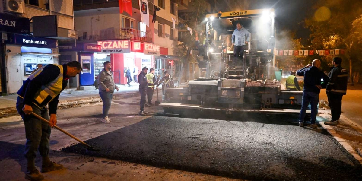 Bornova'da gece saatlerinde bir cadde üzerinde asfalt serme çalışması yapılıyor. Sarı reflektörlü yelek giymiş bir işçi elindeki kürekle asfaltı düzeltirken, arkasında büyük bir Vögele marka asfalt serim makinesi ve diğer görevliler yer alıyor. Arka planda ise ışıkları yanan bir eczane ve bankamatiklerin bulunduğu dükkanlar görülüyor.