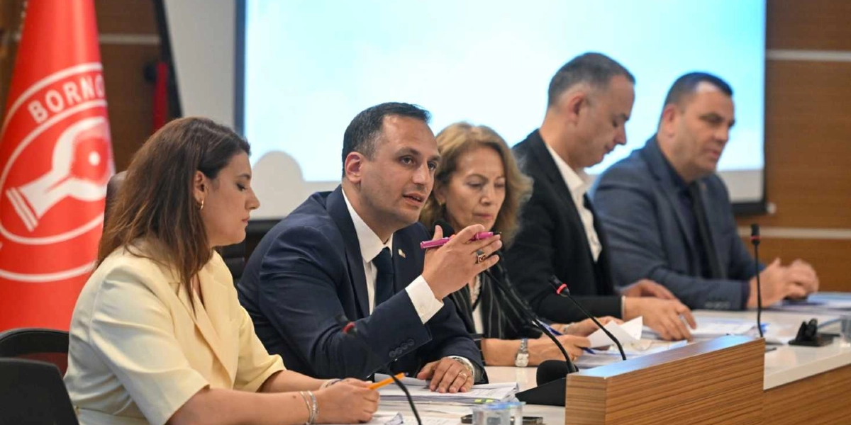 Toplantı masasında yan yana oturan dört kişi, ciddiyetle bir sunumu veya tartışmayı takip ediyor. Masanın en önünde, elinde pembe bir kalemle konuşan bir erkek yer alıyor. Yanında sarı ceketli bir kadın, onun yanında ise koyu renkli kıyafetli bir kadın ve iki erkek daha bulunuyor. Arka planda Bornova Belediyesi'nin kırmızı renkli logosunun yer aldığı bir bayrak ve üzerinde sunum yansıtılan bir ekran görülüyor.