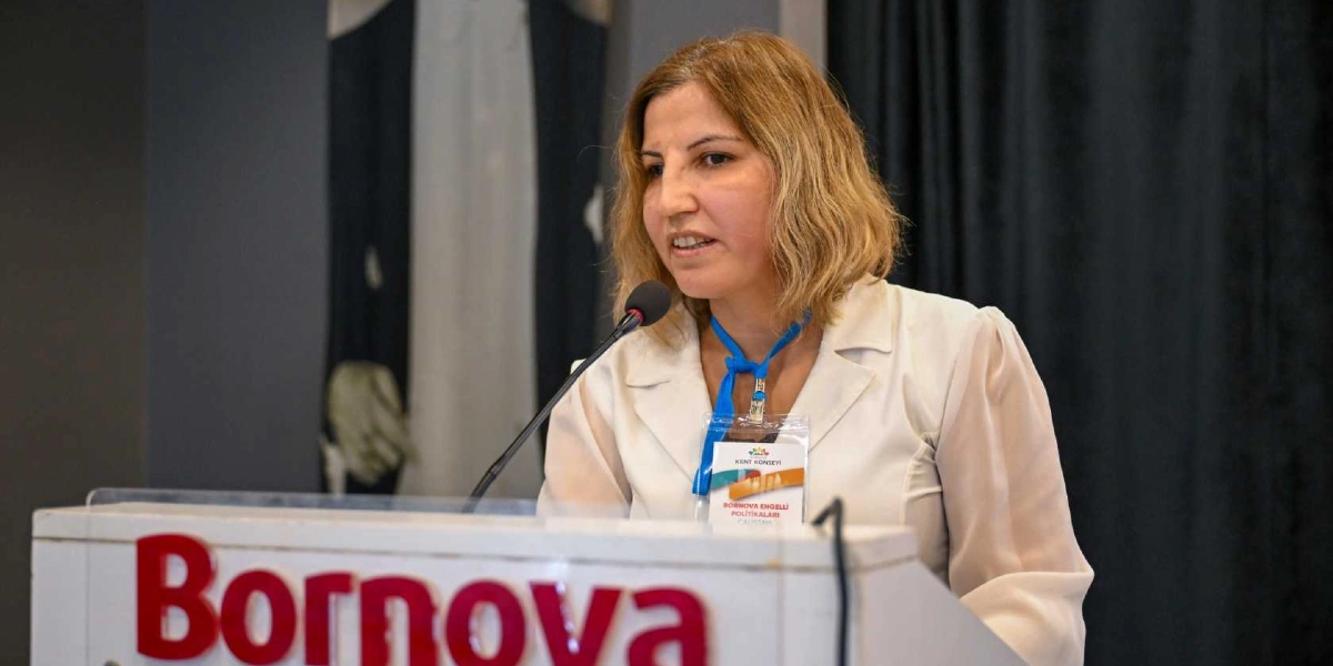 Beyaz ceketli, omuz hizasında sarı saçlı bir kadın, üzerinde Bornova yazılı bir kürsüde mikrofon başında konuşma yapıyor. Boynunda mavi bir yaka kartı asılı olan kadının kartının üzerinde Bornova Kent Konseyi Engelli Politikaları Çalıştayı yazısı okunuyor.