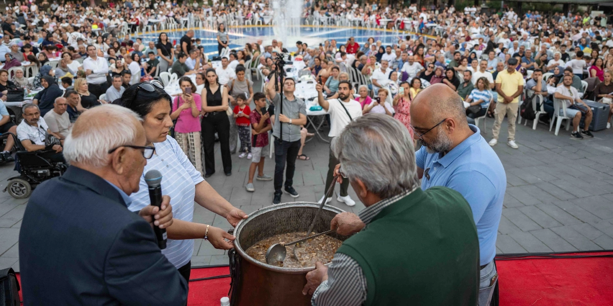 Açık havada, bir meydanda kurulan uzun masalarda oturan kalabalık bir topluluğun önünde, büyük bir kazanda yemek dağıtımı yapılıyor. Ön planda, ellerinde kepçelerle büyük metal kazandan yemek servis eden dört kişi bulunuyor; bunlardan biri elinde mikrofonla konuşuyor. Arka planda ise fıskiyeli bir havuzun etrafına dizilmiş masalarda oturan, etkinliği takip eden çok sayıda insan yer alıyor.
