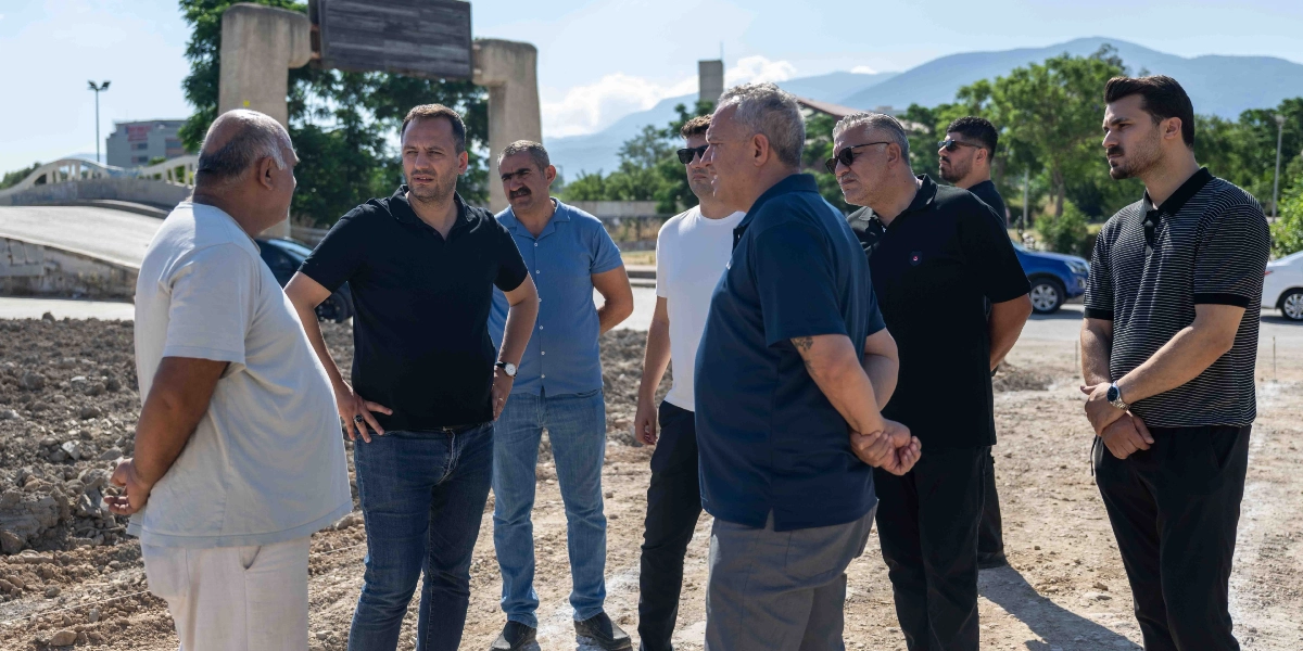 Belediye Başkanı Filiz Ceritoğlu Sengel'in de aralarında bulunduğu bir grup erkek, açık havada, toprak bir zemin üzerinde incelemelerde bulunuyor. Başkan Eşki, yanındaki kişilerle birlikte şantiye alanına benzer bir arazide durarak çevreyi gözlemliyor ve sohbet ediyor. Arka planda ağaçlar, bir köprü girişi ve dağ manzarası yer alıyor.