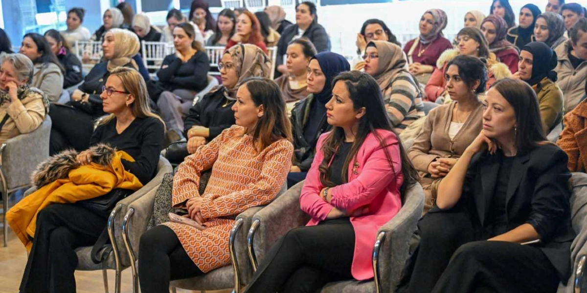Kapalı bir salonda düzenlenen toplantıda, dikkatle dinleyen ve büyük çoğunluğu kadınlardan oluşan kalabalık bir dinleyici grubu sandalyelerde oturuyor. Ön planda, farklı renklerde kıyafetler giymiş kadınlar yan yana dizilmiş sandalyelerde otururken, arka planda daha fazla katılımcı yer alıyor. Katılımcıların birçoğu başörtülü olup, genel atmosfer ciddi ve odaklanmış bir şekilde bir sunumu veya konuşmayı takip ettiklerini gösteriyor.