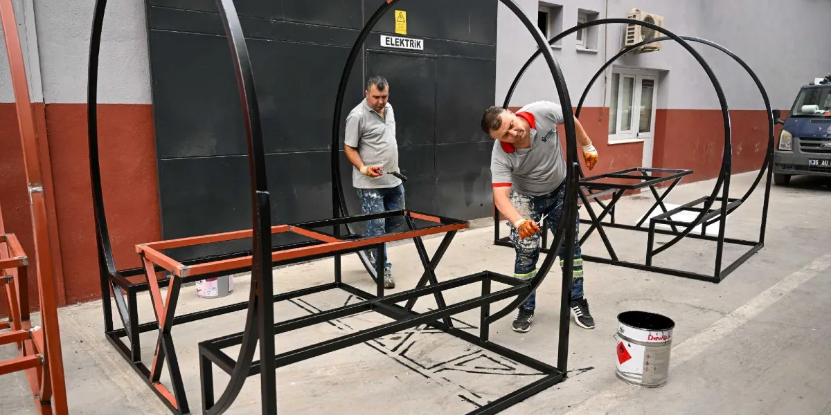 Atölye ortamında iki işçi, parklar için üretilen dairesel metal iskeletli oturma gruplarını siyah boya ile boyuyor. İşçilerden biri elindeki rulo fırça ile metal yapının kavisli kısmını boyarken, diğeri arka planda çalışmaya devam ediyor. Yanlarında boya kutuları ve tamamlanmış diğer oturma üniteleri bulunuyor.