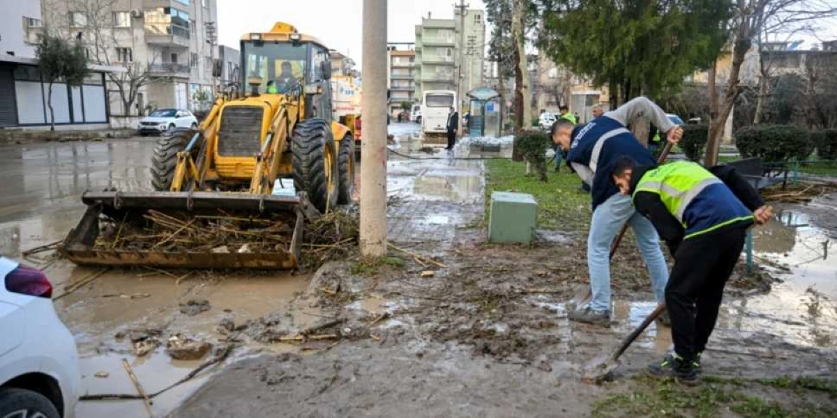 Belediye ekipleri, Bornova'da sel sonrası çamurla kaplanan bir sokakta temizlik çalışması yapıyor. Sarı bir iş makinesi kepçesiyle çamur ve atıkları toplarken, yelekli görevliler küreklerle kaldırım ve yol kenarındaki balçığı temizliyor.