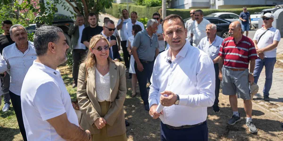Açık havada, yeşil bir alanda bir araya gelmiş kalabalık bir grup insan. Ön planda beyaz gömlekli bir erkek, elinde bir kağıt tutarak konuşuyor; yanında bej renkli bir ceket giymiş güneş gözlüklü bir kadın ve beyaz polo yaka tişörtlü başka bir erkek onu dinliyor. Arka planda ise farklı yaş ve giyim tarzlarında, çoğunluğu erkeklerden oluşan bir grup insan daha bulunuyor.