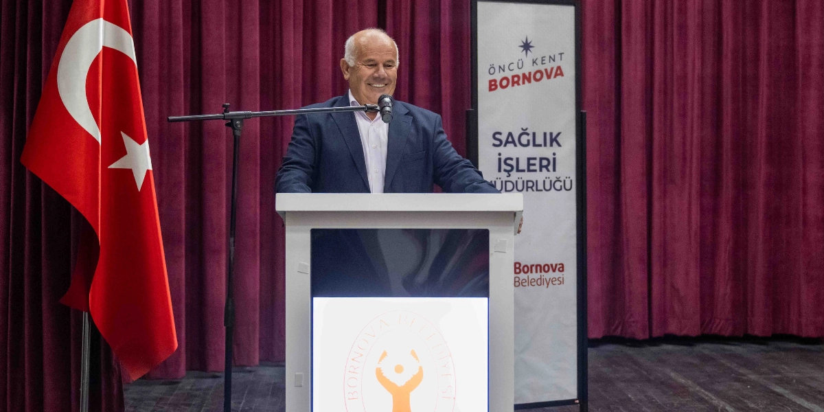 Kürsüde konuşma yapan, beyaz saçlı ve güler yüzlü bir erkek, arkasında bordo renkli perde ve yanında Türk bayrağı ile duruyor. Kürsünün ön yüzünde Bornova Belediyesi logosu yer alırken, sağ taraftaki dikey tabelada 