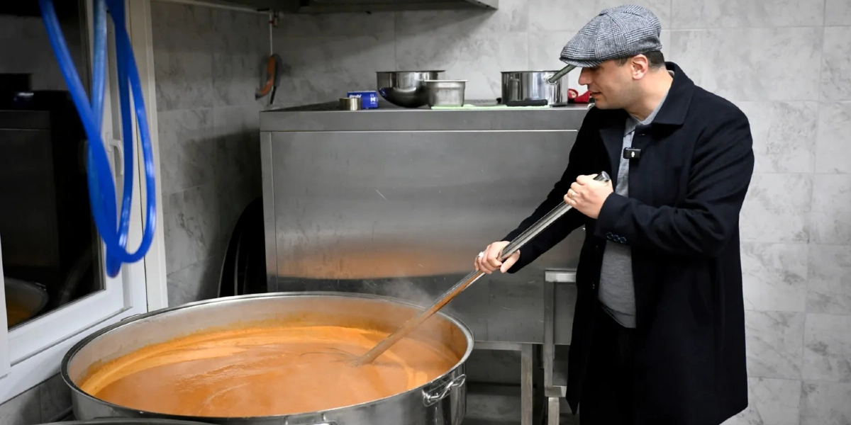 Gri kasketli ve lacivert paltolu bir erkek, endüstriyel bir mutfakta büyük bir metal kazanda pişmekte olan turuncu renkli yemeği uzun bir kepçeyle karıştırıyor.