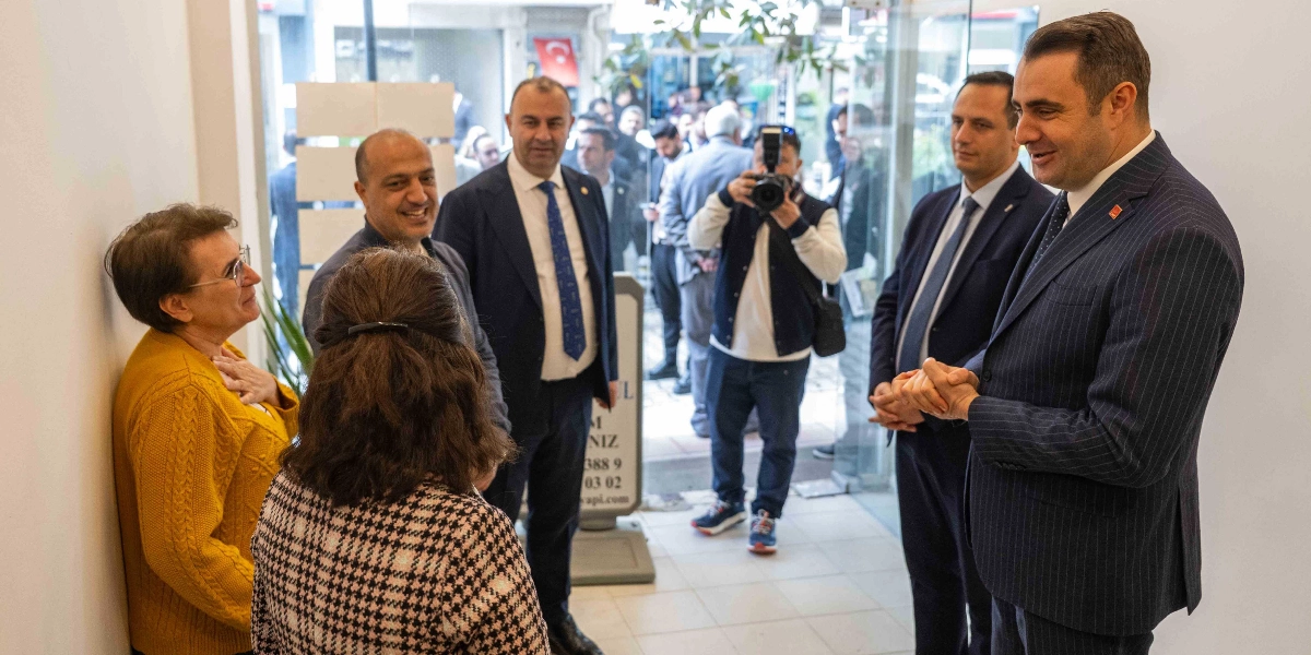 Kapalı bir mekanda, takım elbiseli siyasetçiler ve vatandaşlar bir arada duruyor. Sağ tarafta lacivert çizgili takım elbiseli bir erkek, ellerini birleştirmiş şekilde karşısındakilerle konuşuyor. Sol tarafta sarı hırkalı bir kadın ve desenli ceketli bir başka kadın onu dinliyor. Arka planda ise elinde fotoğraf makinesi olan bir kişi ve diğer katılımcılar yer alıyor.