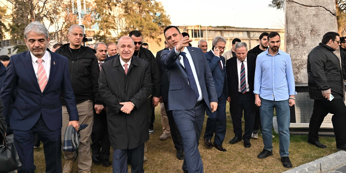 Ümit Özdağ ve beraberindeki bir grup, Abide-i Hürriyet Meydanı'nda yürürken görülüyor. Özdağ koyu renkli bir palto giymiş, ellerini önünde birleştirmiş şekilde ilerliyor. Yanındaki takım elbiseli bir kişi eliyle çevreyi işaret ederek bir şeyler anlatıyor. Grubun sağ tarafında üzerinde yazılar bulunan dikey bir anıt taşı yer alıyor. Arka planda ağaçlar ve meydanın diğer yapıları seçiliyor.