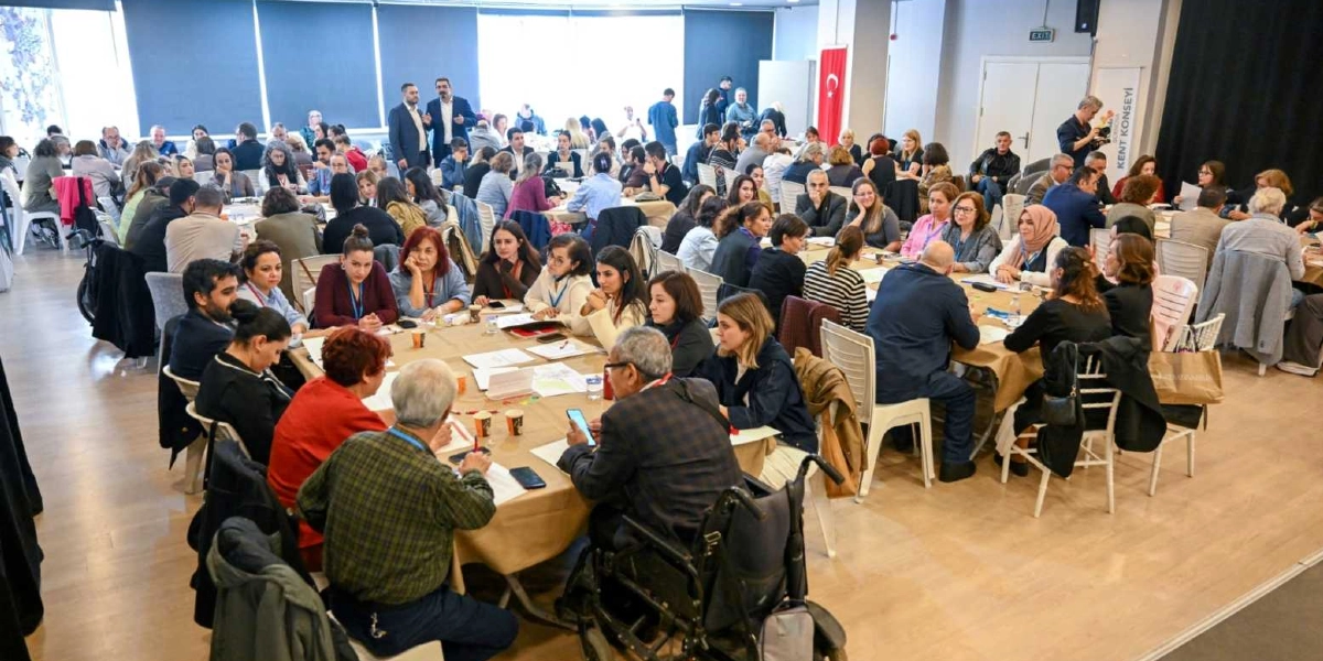 Geniş bir toplantı salonunda yuvarlak masalar etrafında toplanmış, farklı yaş ve kesimlerden çok sayıda katılımcının yer aldığı bir çalıştay. Katılımcılar masalarda oturarak notlar alıyor, birbirleriyle konuşuyor ve fikir alışverişinde bulunuyor. Ön planda tekerlekli sandalye kullanan bir katılımcı da dahil olmak üzere çeşitli bireylerin yer aldığı kapsayıcı bir ortam görülüyor. Arka planda Türk bayrağı ve Bornova Kent Konseyi yazılı bir afiş bulunuyor.