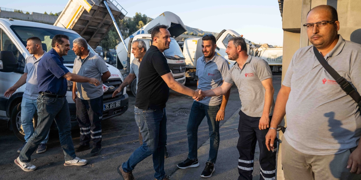 Bornova Belediye Başkanı Ömer Eşki, sabahın erken saatlerinde belediye şantiyesinde çalışan işçilerle bir araya gelerek tokalaşıyor ve sohbet ediyor. Arka planda belediyeye ait çöp toplama araçları ve kamyonlar bulunurken, ön planda üzerinde Bornova Belediyesi logolu gri tişörtler giyen personel yer alıyor.