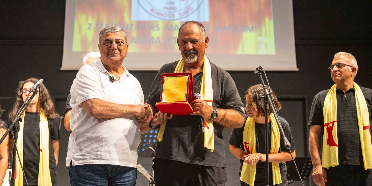Kapalı bir sahnede düzenlenen anma etkinliğinde, beyaz gömlekli bir kişi, üzerinde kırmızı bir plaket bulunan siyah tişörtlü bir adama plaketini takdim ederken el sıkışıyor. Her iki kişinin boynunda üzerinde kırmızı figürler bulunan sarı şallar asılı. Sahnedeki diğer katılımcılar da aynı şallardan takıyor ve arkadaki projeksiyon perdesinde Sivas Madımak Katliamı ile ilgili bir yazı yer alıyor.