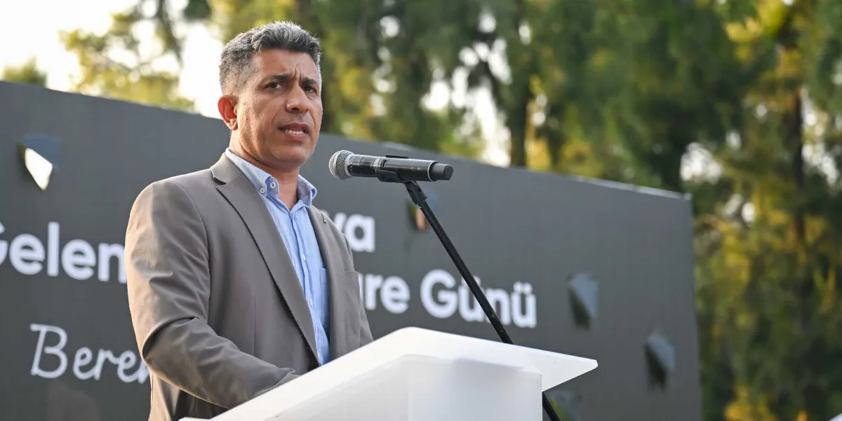 Kısa saçlı, gri ceket ve açık mavi gömlek giyen bir erkek, dış mekanda beyaz bir kürsüde mikrofon başında konuşma yapıyor. Arkasındaki koyu renkli panoda 