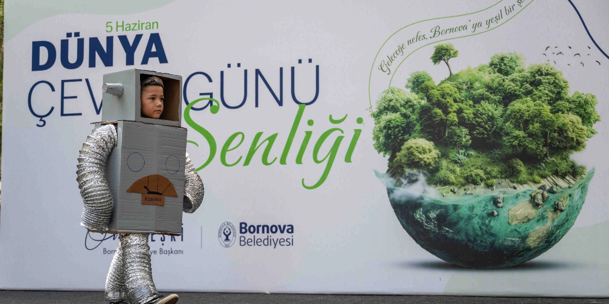 Karton kutulardan yapılmış robot kostümü giymiş bir çocuk, arkasında 5 Haziran Dünya Çevre Günü Şenliği yazılı büyük bir afişin önünde yürüyor. Afişin sağ tarafında yeşil bir orman ve dünya küresi görseli ile üzerinde 