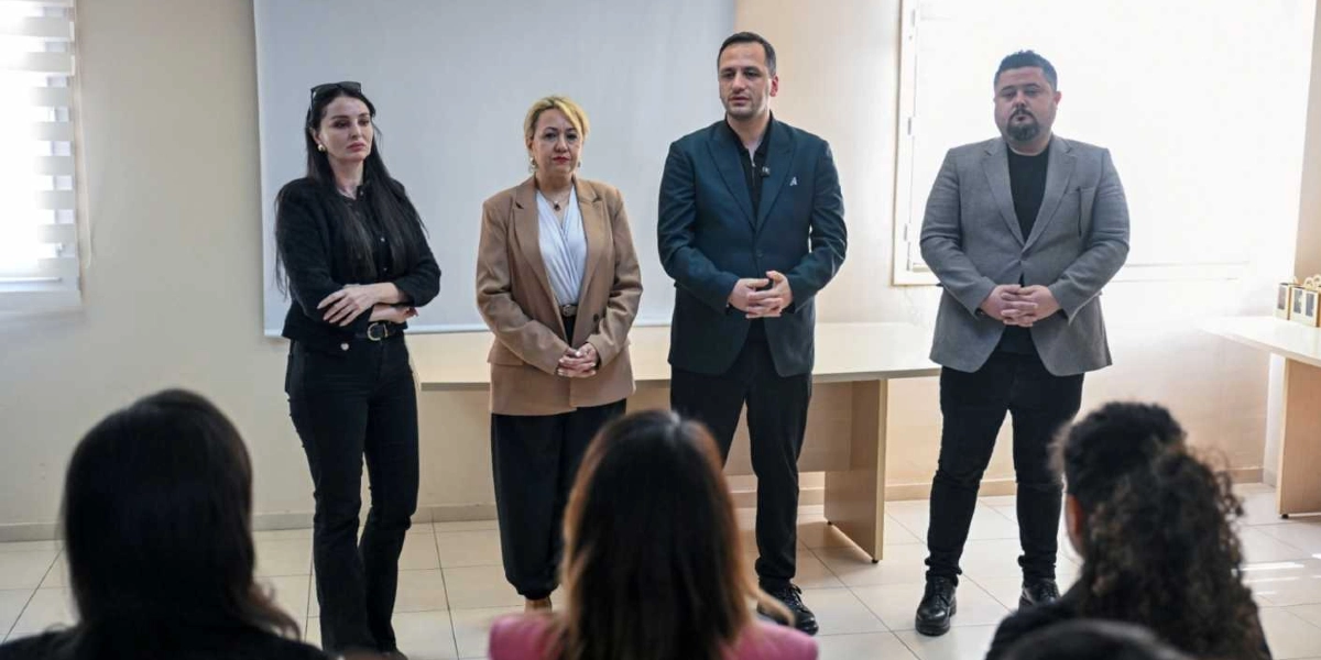 İç mekanda, beyaz bir duvarın önünde yan yana duran iki kadın ve iki erkekten oluşan bir grup, karşılarındaki katılımcılara hitap ediyor. Soldan sağa doğru; siyah kıyafetli bir kadın, bej ceketli bir kadın, koyu renk ceketli bir erkek ve gri ceketli bir erkek yer alıyor. Ön planda, toplantıyı dinleyen kişilerin başlarının arkası görülüyor.