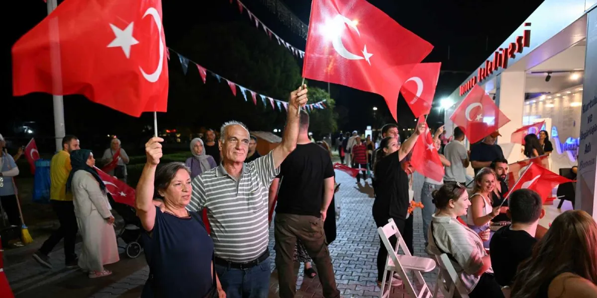 Gece vakti açık havada düzenlenen bir etkinlikte, ellerinde Türk bayrakları tutan insanlar Bornova Belediyesi standının önünde bir arada bulunuyor. Ön planda çizgili tişörtlü bir adam ve yanında lacivert bluzlu bir kadın ellerinde bayraklarla gülümsüyor. Arka planda ise standı ziyaret eden kalabalık bir grup ve ışıklandırılmış stant yapısı görülüyor.