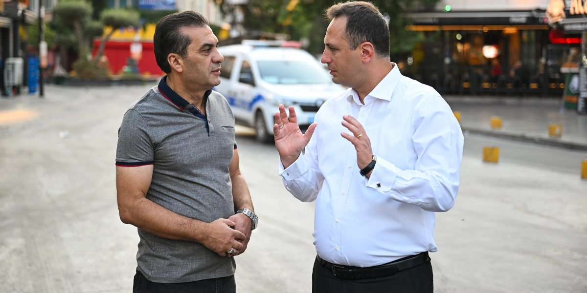 Bornova Belediye Başkanı Ömer Eşki, beyaz gömlekli ve siyah pantolonlu haliyle, gri polo yaka tişört giyen bir kişiyle açık havada sohbet ediyor. Arka planda park halindeki bir resmi araç ve işletmelerin bulunduğu bir cadde yer alıyor.