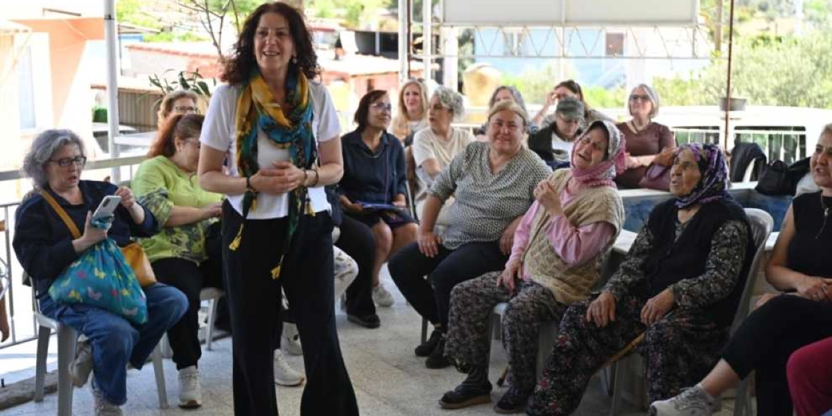 Açık havada, yarı kapalı bir alanda toplanmış bir grup kadın. Ön planda, beyaz tişörtlü ve renkli şallı bir kadın ayakta konuşma yaparken, arkasında ve yanlarında sandalyelerde oturan farklı yaş gruplarından kadınlar onu dinliyor. Katılımcıların bir kısmı başörtülü, bir kısmı ise günlük kıyafetlerle oturuyor ve ortamda samimi, katılımcı bir atmosfer hakim.