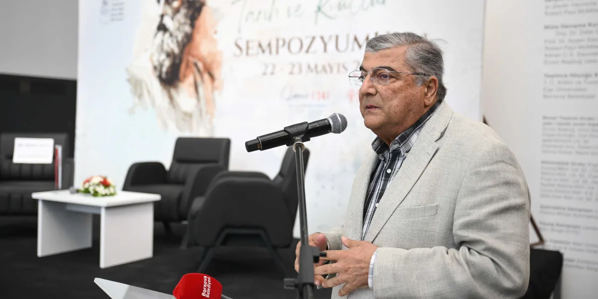 Gri ceketli ve gözlüklü bir konuşmacı, kürsüdeki mikrofon önünde hitap ederken arka planda üzerinde 
