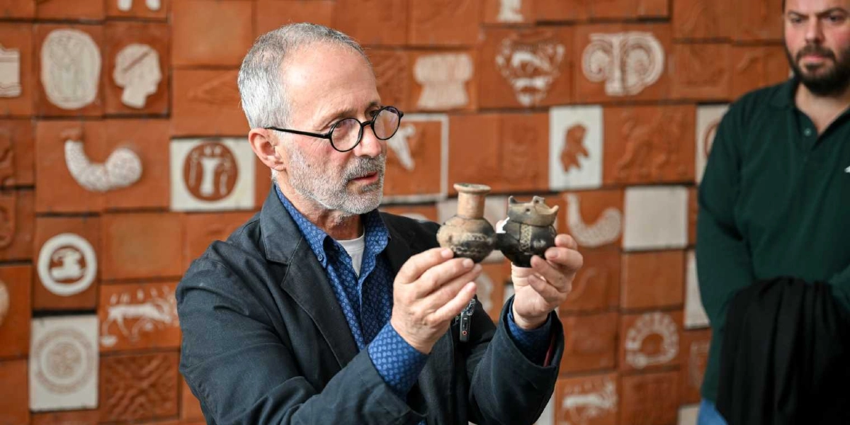 Gözlüklü, gri saçlı ve sakallı bir arkeolog, üzerinde antik desenler bulunan küçük, çift hazneli pişmiş toprak bir eseri elleriyle tutarak inceliyor. Arka planda, üzerinde çeşitli figürlerin ve sembollerin kabartma olarak işlendiği pişmiş toprak karolarla kaplı bir duvar yer alıyor. Sağ tarafta ise bu kişiyi izleyen sakallı bir erkek daha bulunuyor.