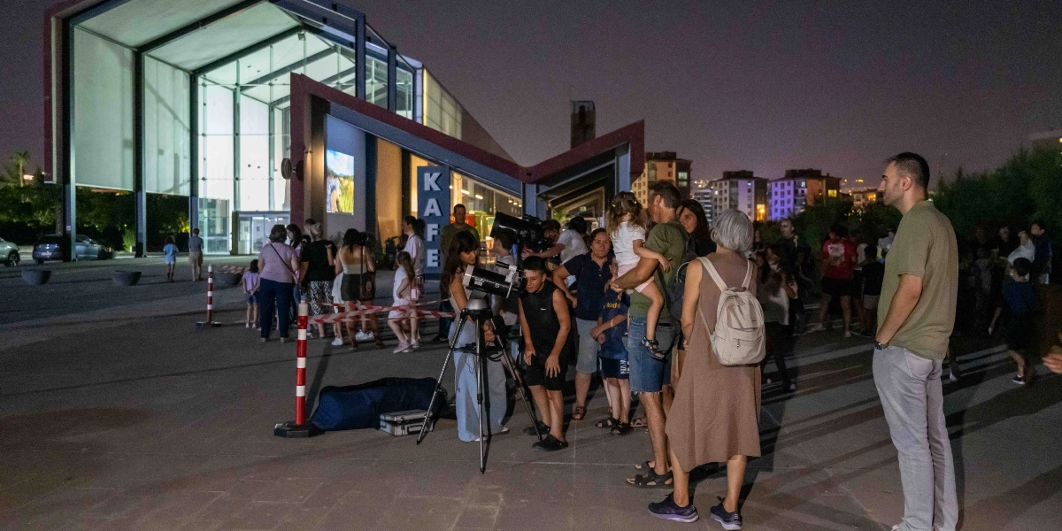 Gece vakti modern bir mimari yapının önünde toplanmış insanlar, tripod üzerine yerleştirilmiş teleskoplarla gökyüzü gözlemi yapıyor. Arka planda 